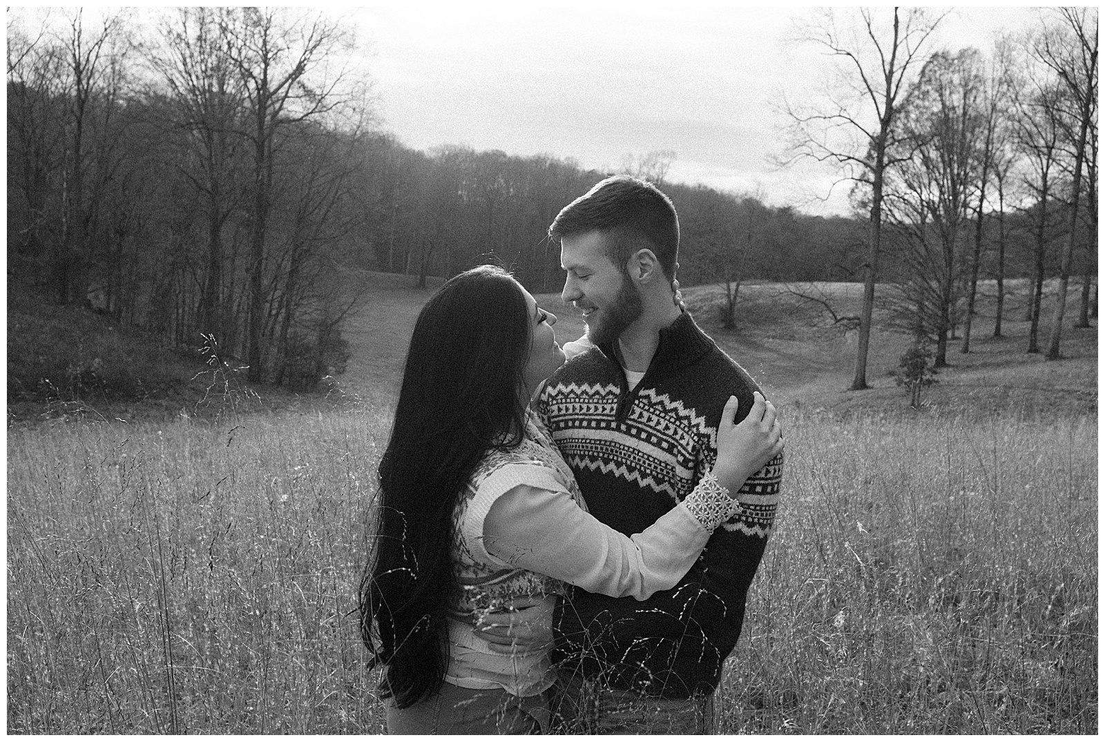 Engaged man and woman snuggled up in meadow at sunset in sweaters for their winter engagement session.