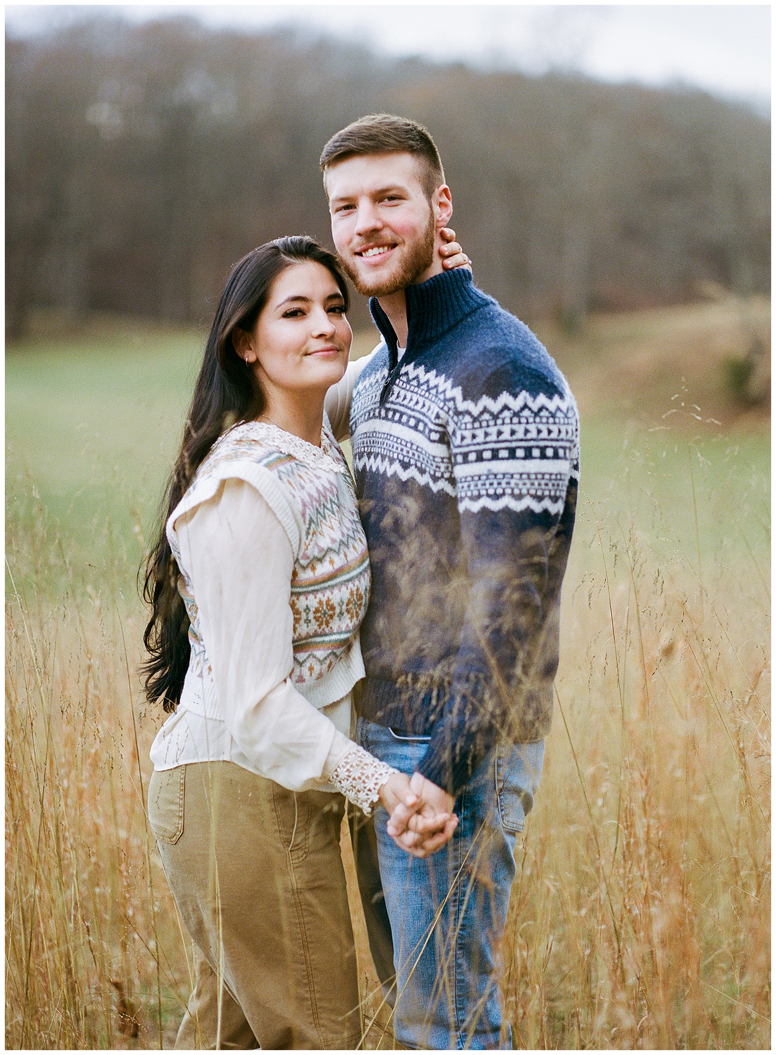 Engaged man and woman snuggled up in meadow at sunset in sweaters for their winter engagement session.