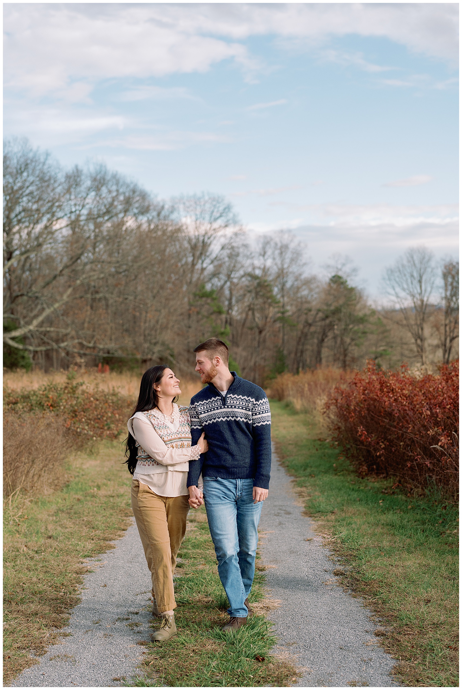 Engaged man and woman wearing sweaters in winter walking holding hands at their farm engagement session.