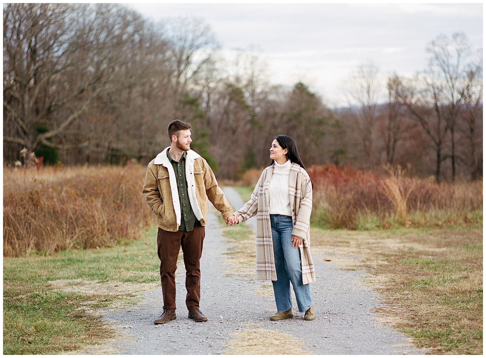 Engaged man and woman wearing winter coats in winter walking holding hands at their farm engagement session.