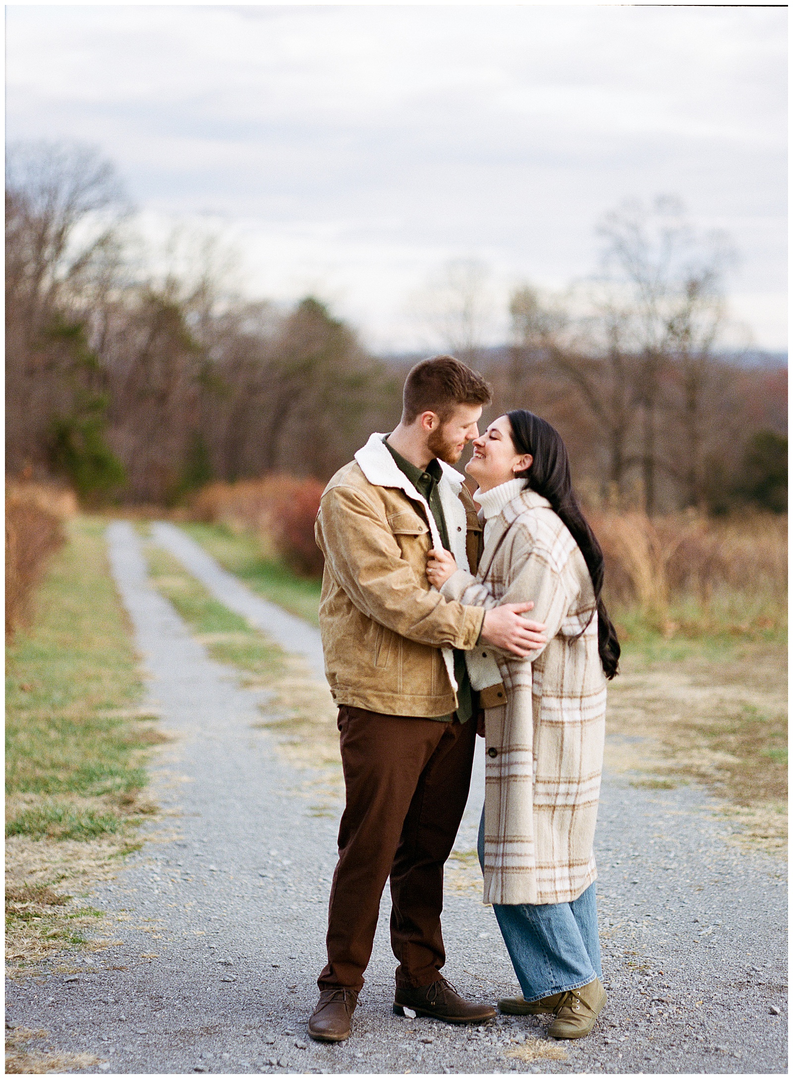 Engaged man and woman wearing winter coats in winter snuggled up at their farm engagement session.