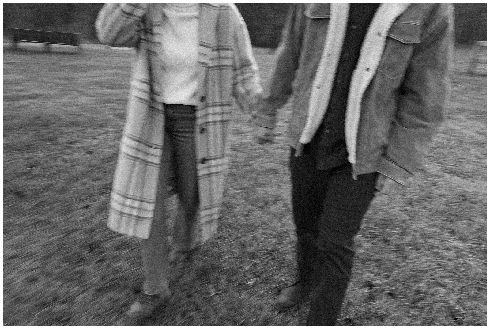 Engaged man and woman wearing winter coats in winter walking holding hands at their farm engagement session; blurry black and white film image
