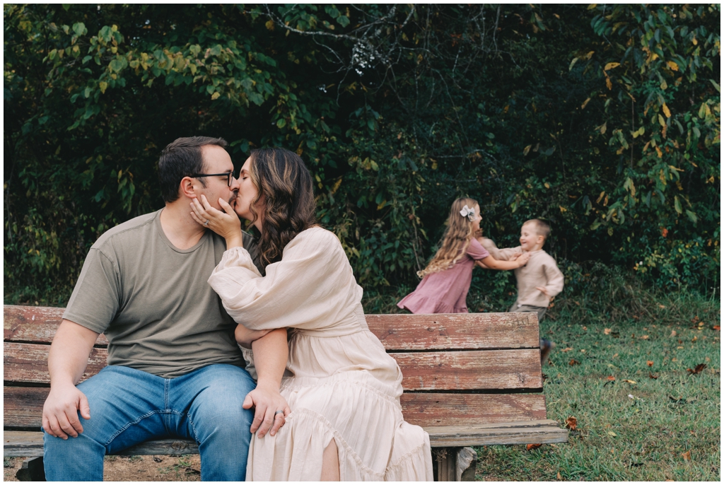 Mom and dad share a kiss while kids play in background during fall family session in Knoxville, TN.