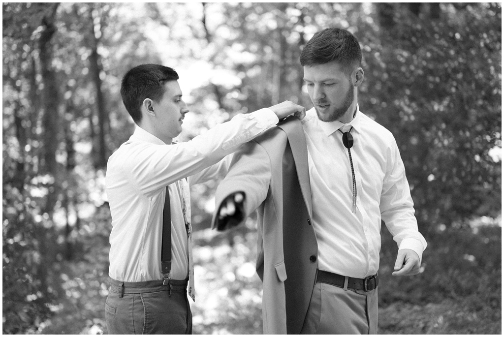 Groomsman helps groom putting on his suit coat in black and white image.