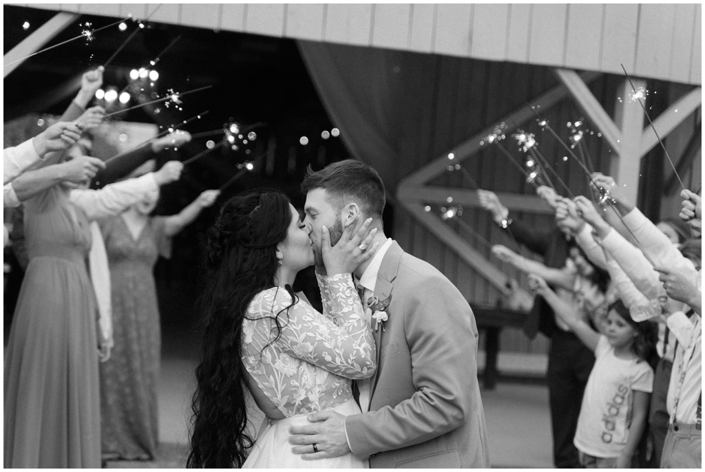 Black and white image of bride and groom kissing while exiting the wedding with sparklers. 