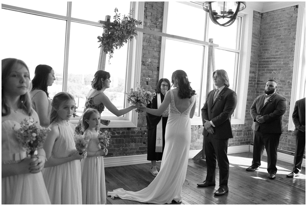 Black and white image of bride passing floral bouquet off to maid of honor during ceremony.